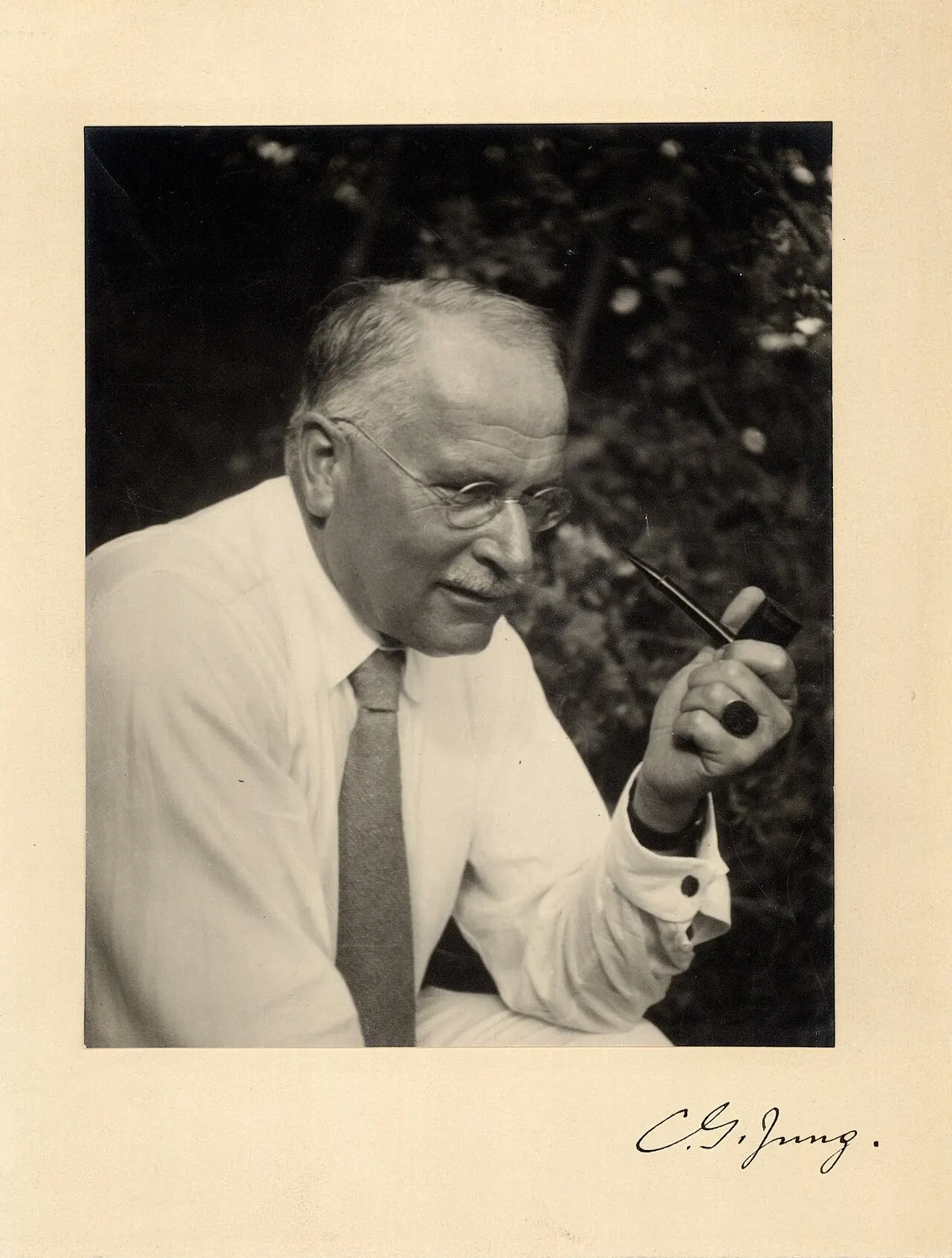 Foto hitam putih Carl Gustav Jung sekitar tahun 1935, berkacamata bulat, berkemeja putih dan berdasi, memegang pipa sambil menunduk, dengan tanda tangan C. G. Jung di bawah foto