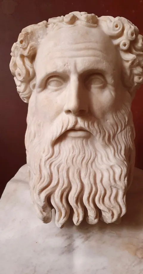Foto patung kepala marmer Zeno dari Kition dengan dahi berkerut, rambut ikal pendek, dan janggut panjang bergelombang, berlatar dinding merah tua di Neues Museum Berlin